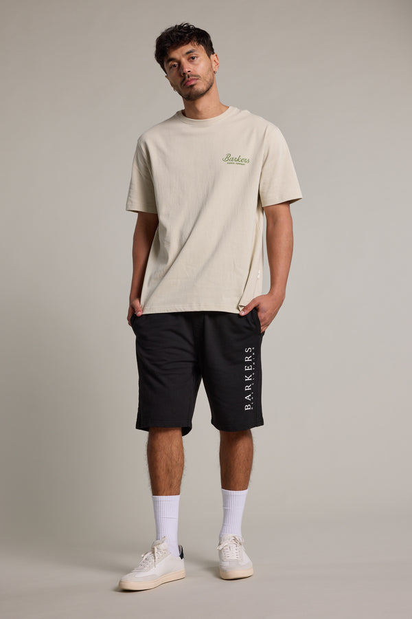 A man stands against a plain background wearing a beige Barkers t-shirt, Organic Classic Print Track Shorts by Barkers in black, white socks, and white sneakers. He has short hair, a trimmed beard, and his hands in his pockets.