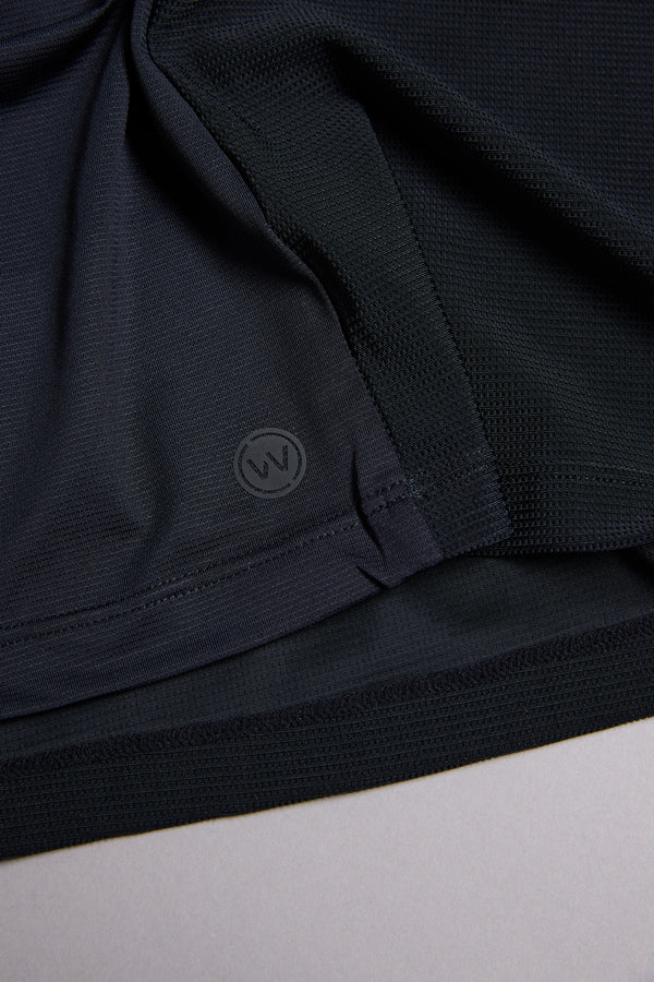 Close-up of the WearEver Pulse Tee's dark, textured performance fabric with a stitched hem and a small circular WearEver "W" logo near the edge, lying flat on a light surface.
