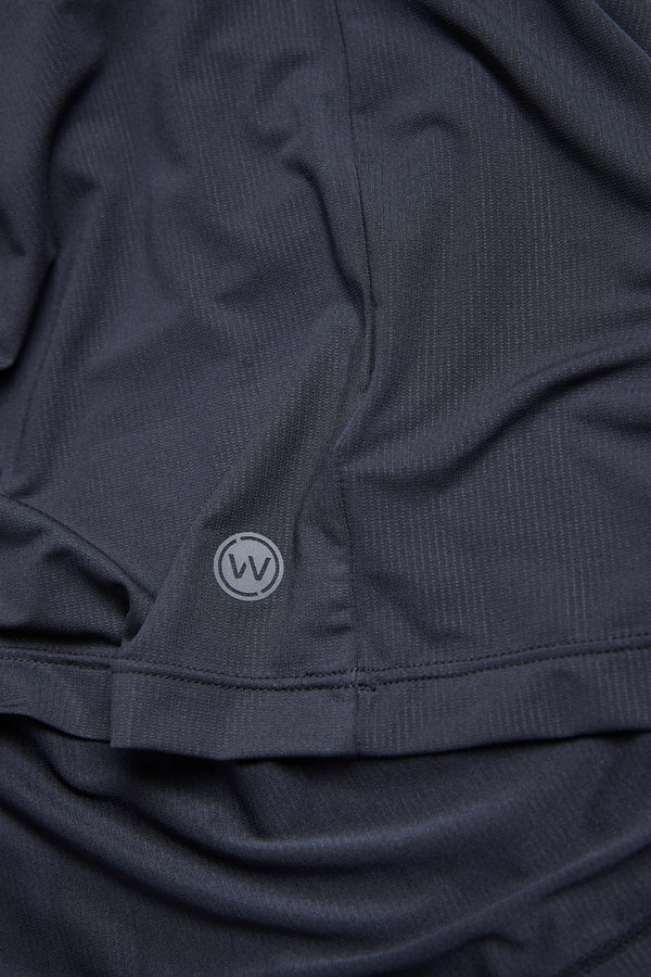 A close-up of the WearEver Pulse Long Sleeve Tee by WearEver in dark gray, featuring a textured fabric, subtle folds, a small circular WV logo near the hem, and UPF protection for sun safety.