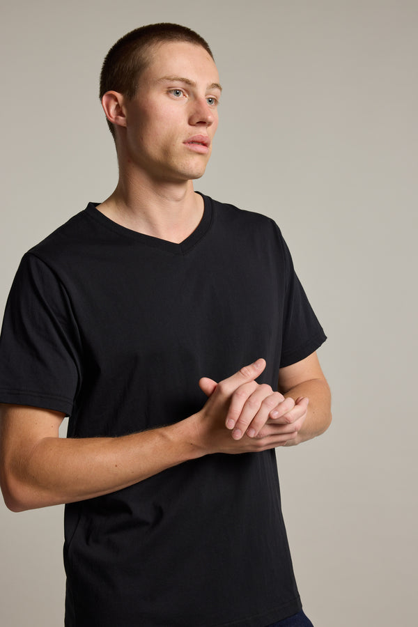 A young man with short hair stands against a plain light background, wearing the Barkers Organic Cotton V Neck Tee—an everyday essential—as he looks to the side with his hands clasped.