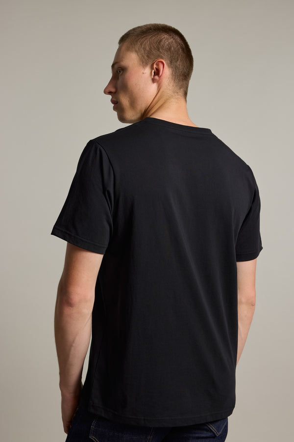 A man with short brown hair, wearing the Barkers Organic Cotton V Neck Tee in black and dark jeans, stands facing away from the camera and looks over his left shoulder, set against a plain, light background.