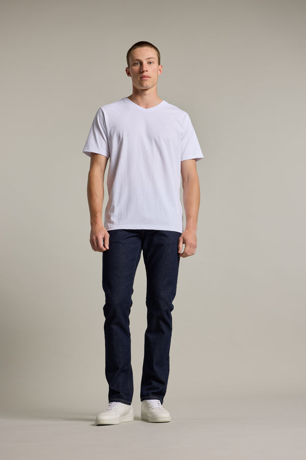 A man with short hair and a neutral expression faces forward against a plain light background, wearing the Barkers Organic Cotton V Neck Tee, paired with dark blue jeans and white sneakers.