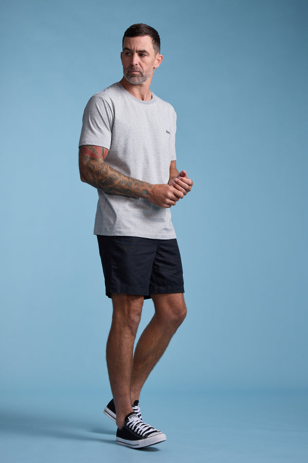A man with short dark hair and tattooed arms stands against a light blue background, wearing the Bayside Emb Tee by Barkers in light gray, black shorts, and black sneakers with white soles, looking slightly to his left.