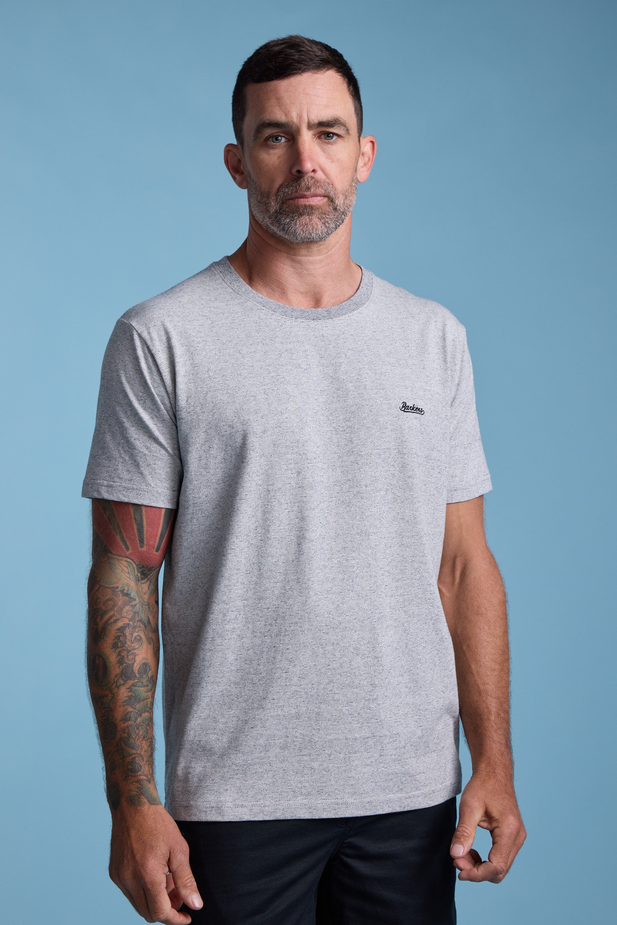 A man with short dark hair, a trimmed beard, and a tattoo sleeve on his left arm wears the Barkers Bayside Emb Tee in light gray and black pants, standing against a plain light blue background.