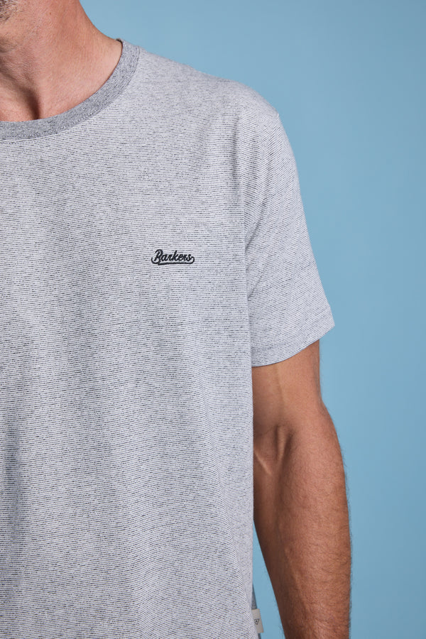 A person wears the Barkers Bayside Emb Tee—light grey, organic cotton, short sleeves with a small black collegiate logo on the left chest. Only the upper torso and part of the left arm show against a blue background.