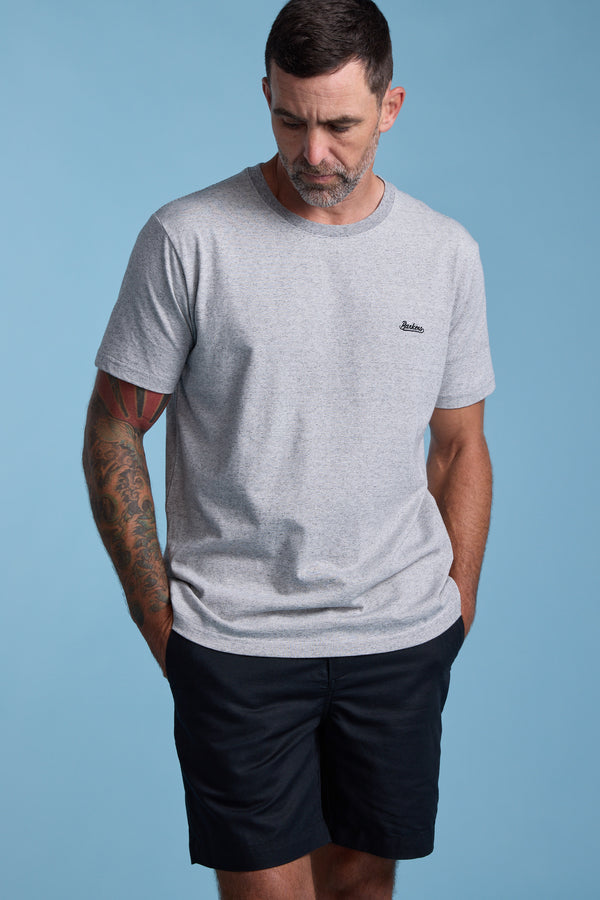 A man with short dark hair and tattoos on his left arm wears the Barkers Bayside Emb Tee, a light gray organic cotton t-shirt, paired with black shorts. He stands against a blue background, hands in pockets, looking down.