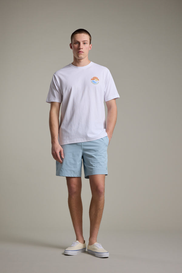 A person stands facing forward, wearing a Barkers Long Board Classic Print Tee with a small chest graphic, light blue shorts, and white shoes, against a plain light gray background.