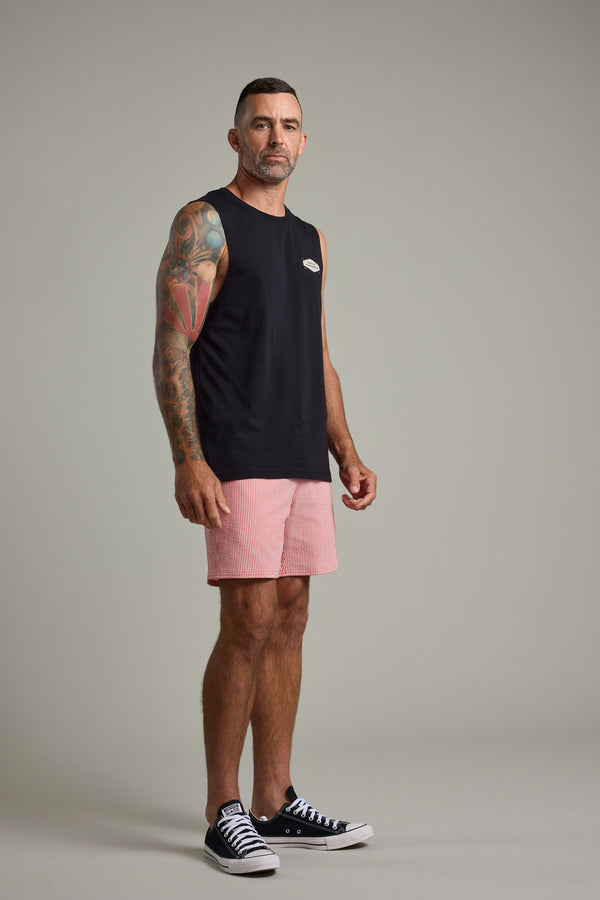 A man with short hair and arm tattoos stands sideways against a light gray background, wearing a black sleeveless shirt, Barkers Dehaven Stripe Volley quick-drying pink swim trunks, and black sneakers.