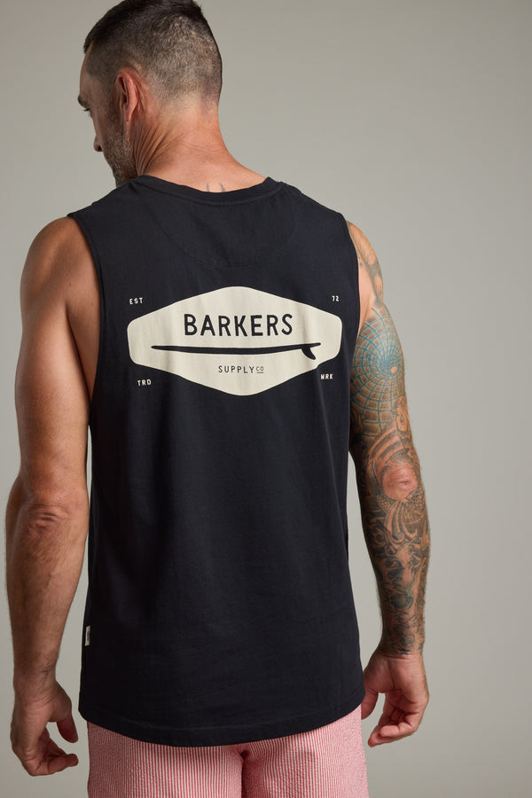 A man with short hair and a tattooed right arm wears the black Barkers Supply Co Print Singlet with logo on the back, facing away from the camera in pink and white striped shorts—a true summer essential by Barkers.