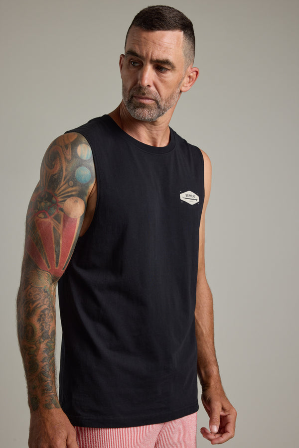 A man with short dark hair and a trimmed beard wears the Barkers Supply Co Print Singlet by Barkers, featuring a small white logo. His colorful tattooed arm stands out as he looks to his left against a plain background—a summer essential.