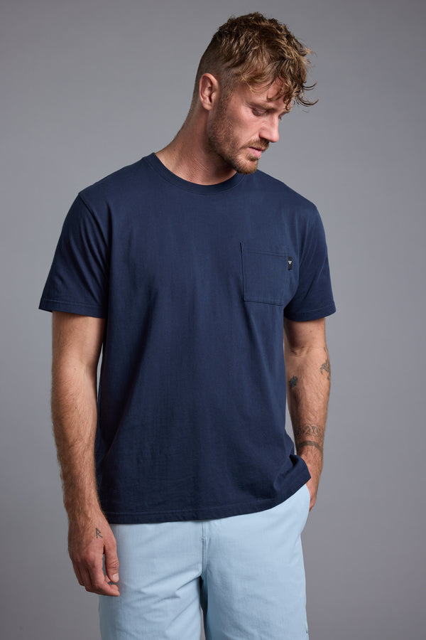 A man with short, messy hair wears a Barkers Summer Soul Sessions Print Tee and light blue pants, standing against a plain gray background, looking down with one hand in his pocket.