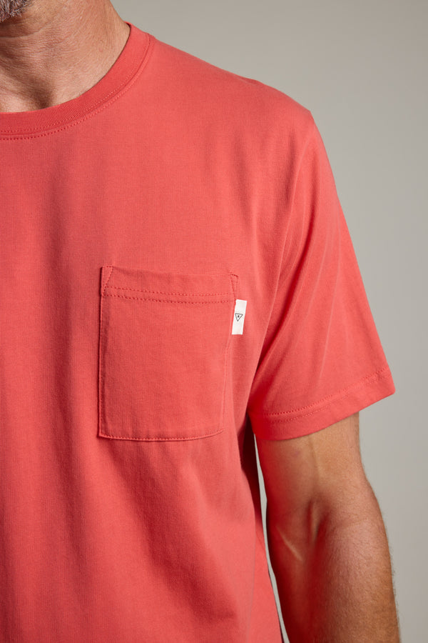 A close-up of a person in Barkers' Summer Soul Sessions Print Tee, showcasing the coral hue, left chest pocket with a white tag, and soft organic cotton. Only the lower face, neck, and upper torso are visible against a neutral backdrop.