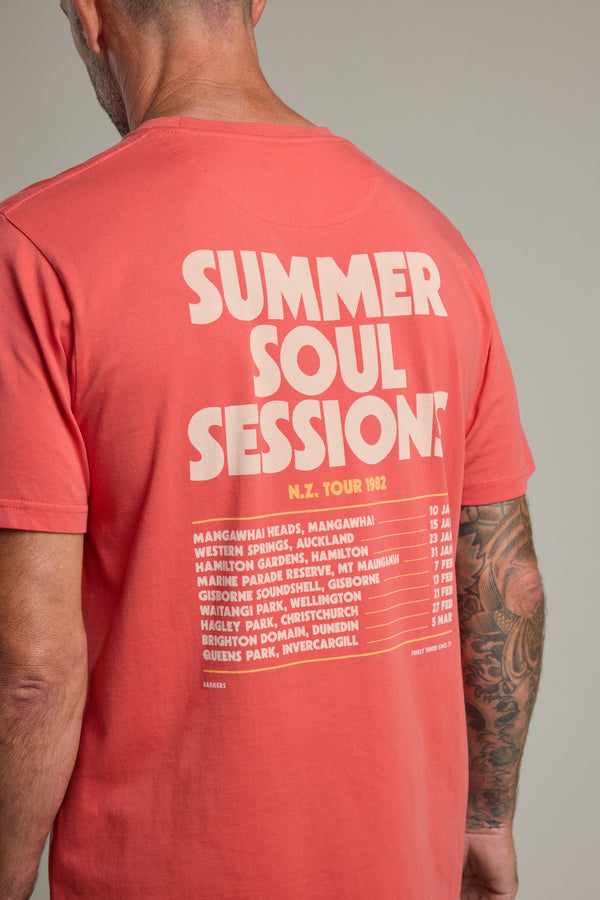 A man wears the Barkers Summer Soul Sessions Print Tee in coral, made from organic cotton, featuring "SUMMER SOUL SESSIONS N.Z. TOUR 1982" and New Zealand tour locations boldly printed in cream on the back.
