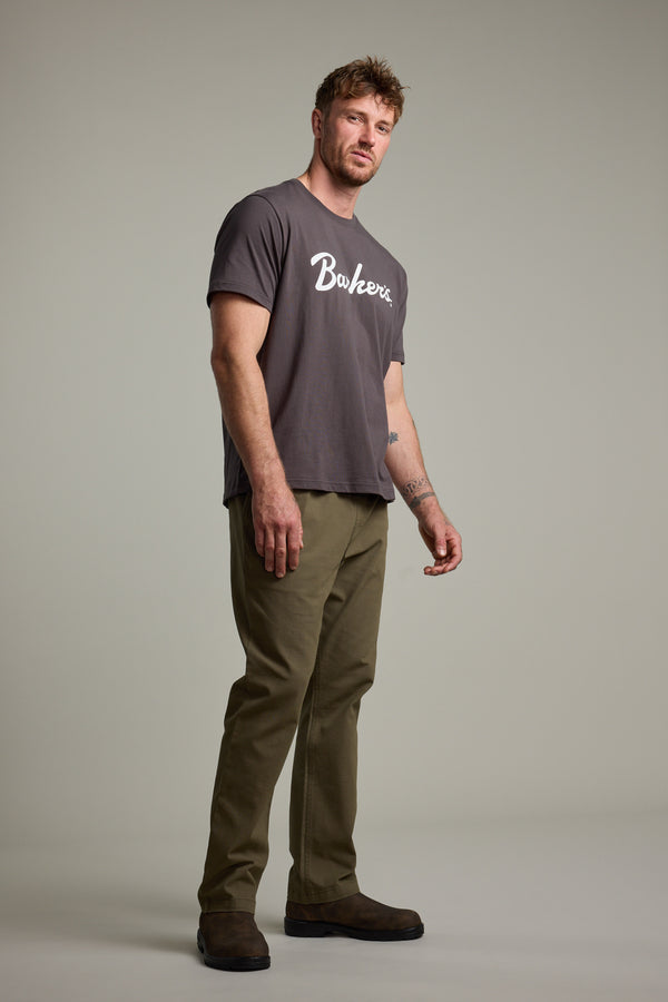 A man with short hair and a light beard stands against a plain background, wearing Barkers’ Finely Tuned Print Tee in dark gray with a vintage crackle logo, paired with olive green pants and brown shoes, looking slightly to the side.