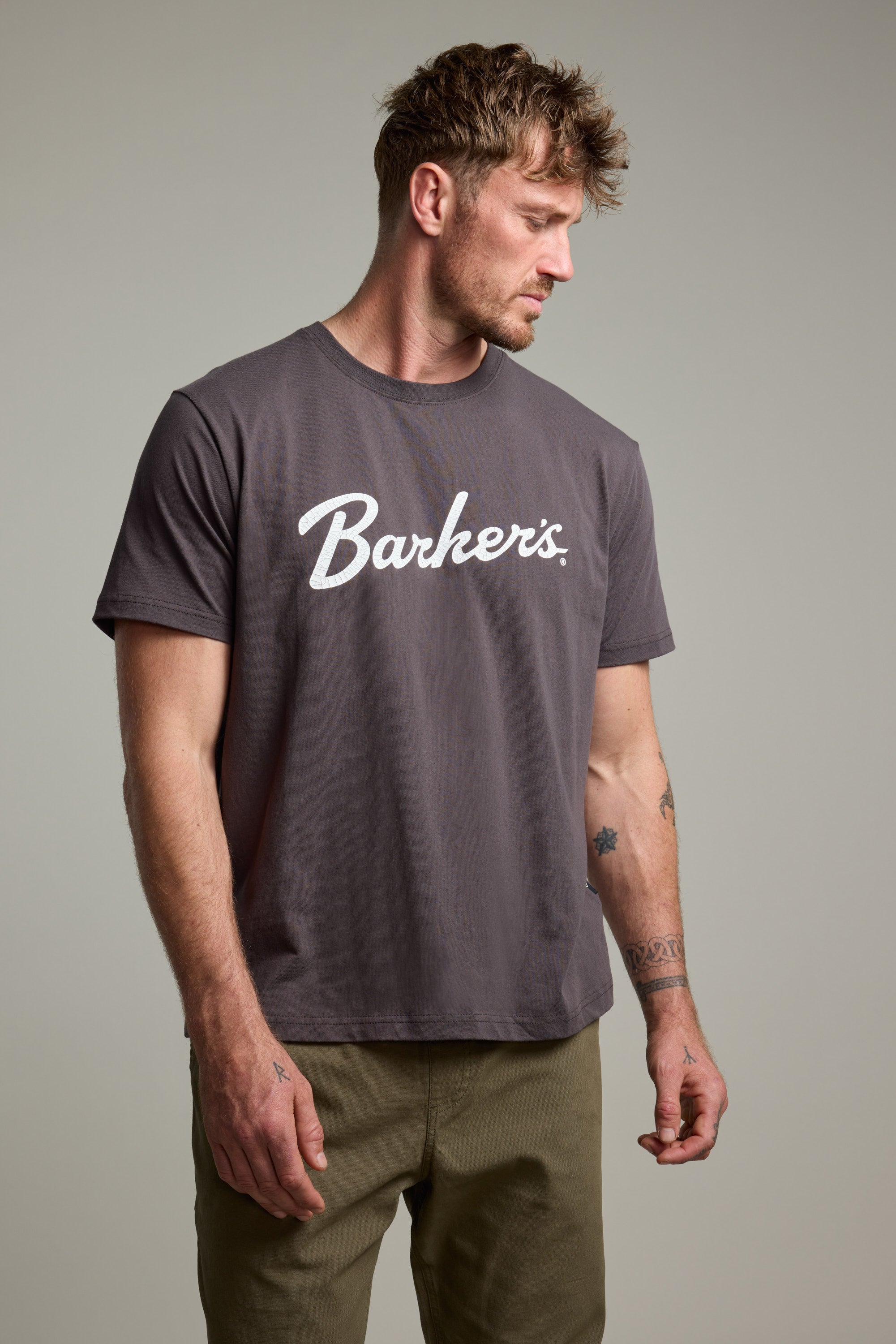 A man with short, tousled hair and visible arm tattoos wears a gray Barkers Finely Tuned Print Tee in organic cotton and olive pants, standing against a plain light background while looking to his left.
