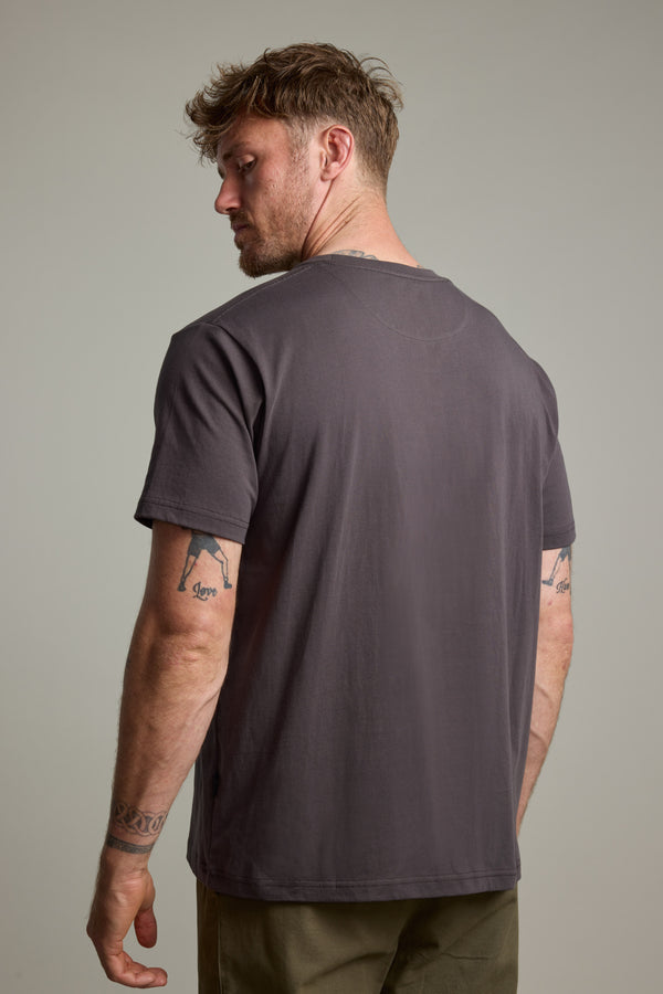 A man with short, tousled hair and visible arm tattoos stands with his back to the camera, wearing a Barkers Finely Tuned Print Tee and olive green pants against a neutral background.