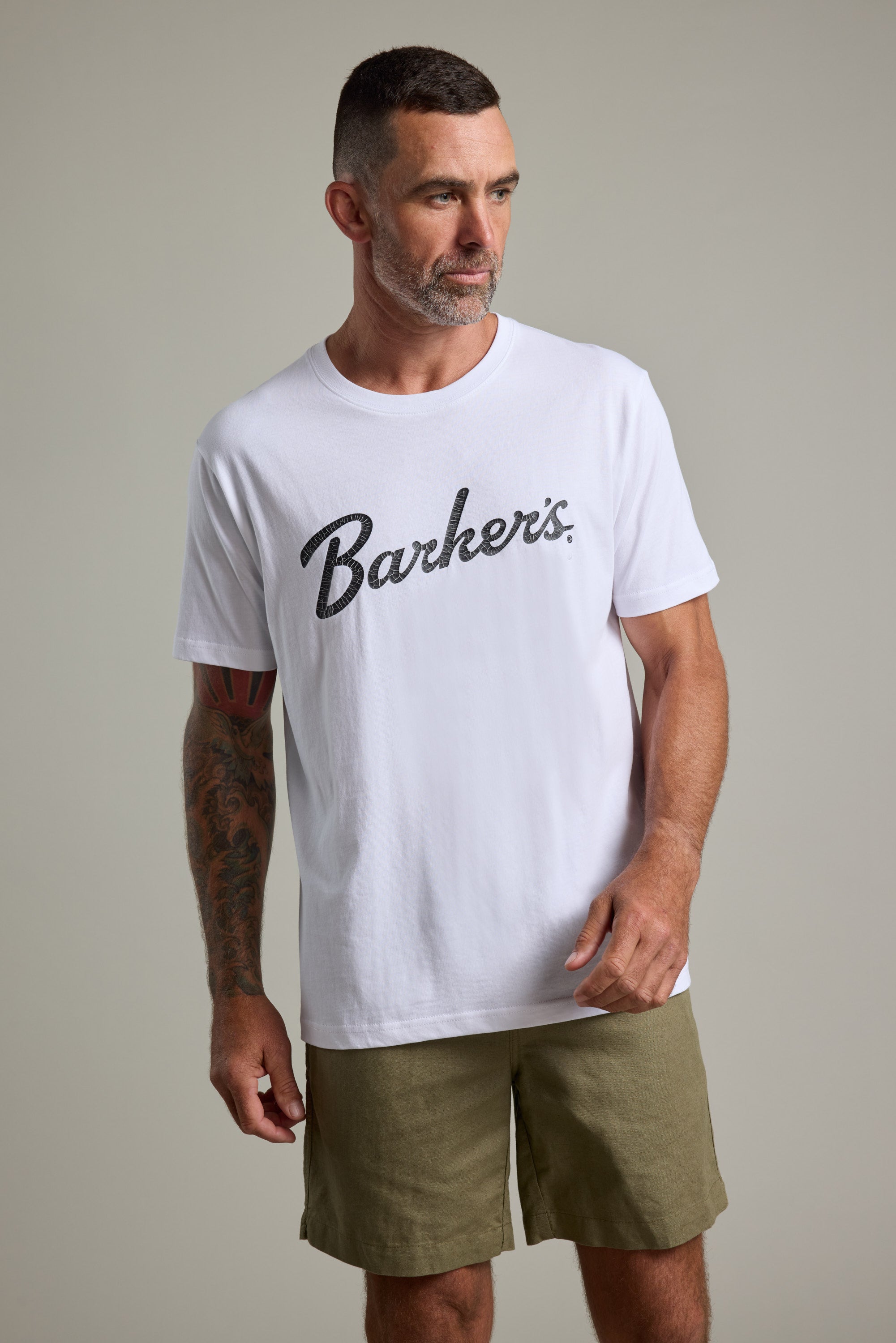A bearded man with short dark hair wears a white Barkers Finely Tuned Print Tee and olive green shorts. He has a tattoo on his left arm and stands against a plain light gray background, looking to his left.