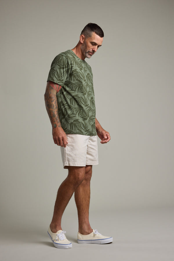 A man with short dark hair and tattoos on his right arm wears a Barkers Palm Leaves Print Tee, light shorts, and white shoes, standing against a plain light-gray background.