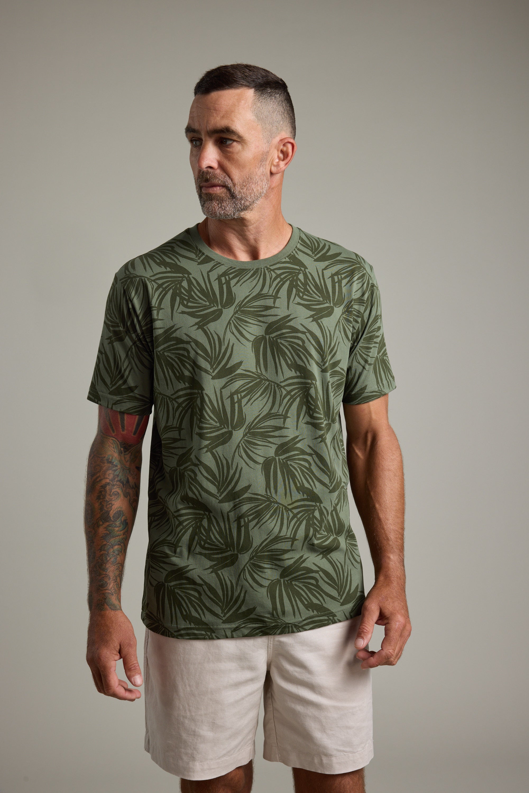 A man with short dark hair and a trimmed beard wears the Barkers Palm Leaves Print Tee and light shorts, standing against a plain gray background. He has a tattoo sleeve on one arm and is looking slightly to his left.
