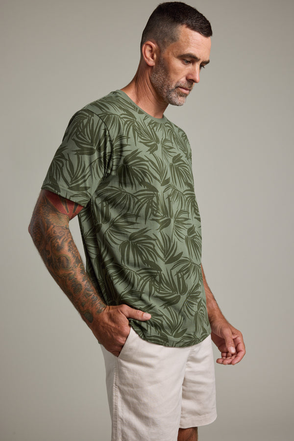 A man with short dark hair and a beard, wearing a Barkers Palm Leaves Print Tee and light shorts, stands with his hands in his pockets looking down against a neutral background.