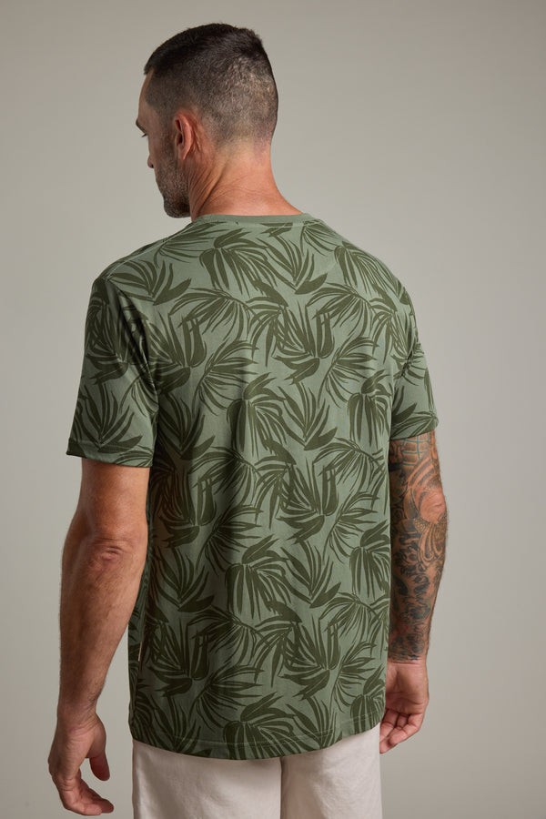 A man with short hair and a beard, sporting a tattoo sleeve, is seen from behind in light pants and the Barkers Palm Leaves Print Tee—an organic cotton shirt featuring a dark palm leaf pattern—against a plain, neutral background.