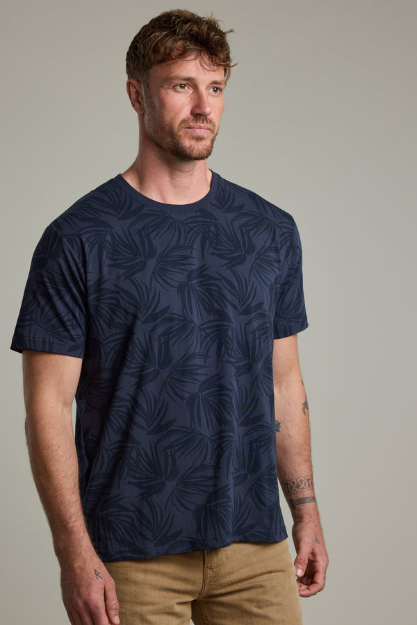 A man with short brown hair and a beard wears the Barkers Palm Leaves Print Tee in navy blue and beige pants, standing against a plain light-gray background and looking slightly to the side.