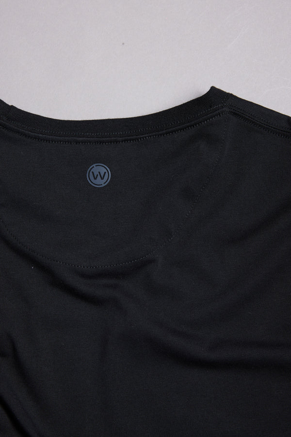 A close-up of the neckline and shoulder of the WearEver Outbound T-Shirt by WearEver, in black cotton stretch fabric, shows a small circular “W” and “V” logo near the back collar. The soft fabric looks smooth and comfortable.