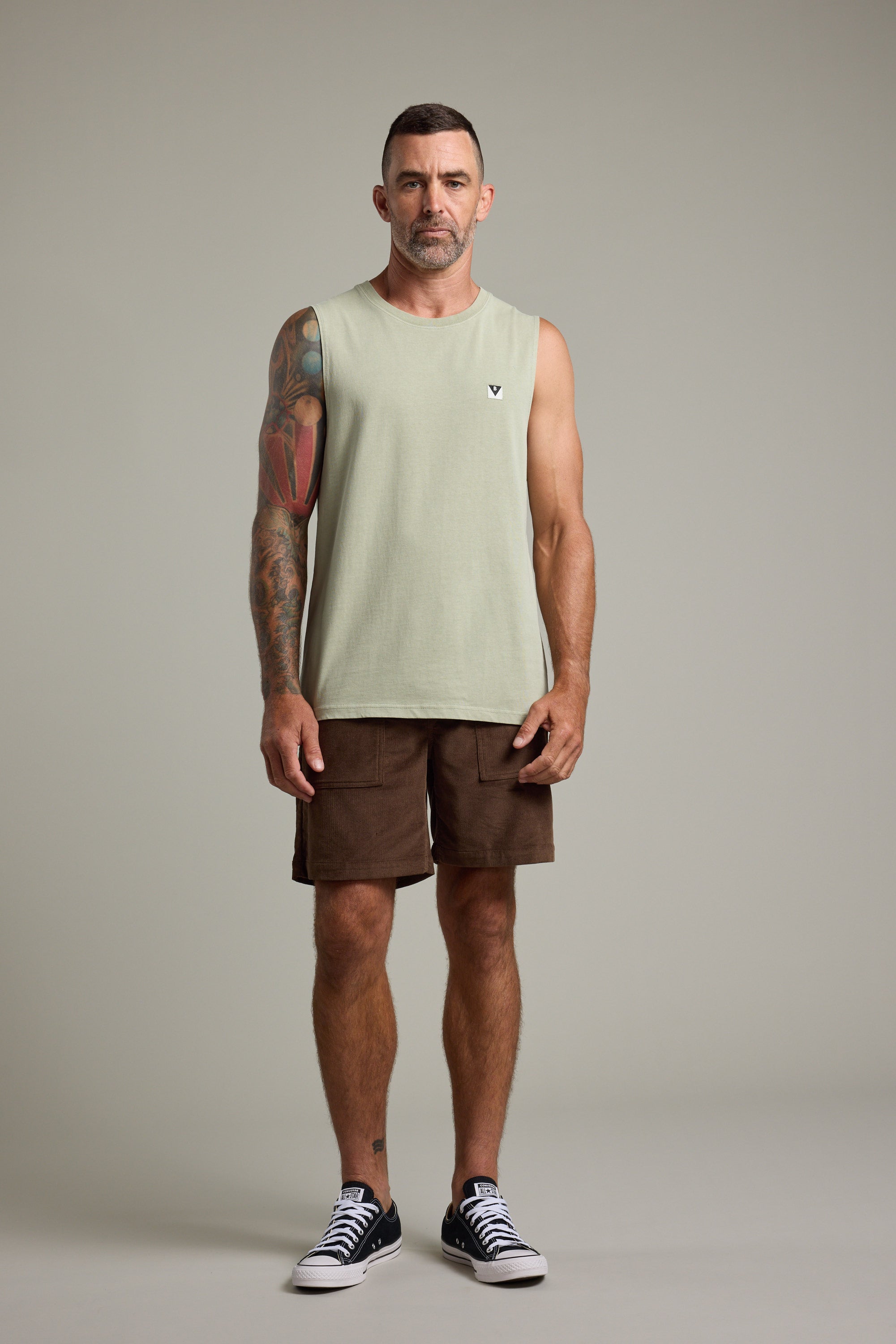 A bearded man with short hair stands facing forward, wearing the Barkers Newton Washed Singlet in light green, brown shorts, and black sneakers. A colorful tattoo sleeve covers his right arm. The plain backdrop adds a vintage feel.