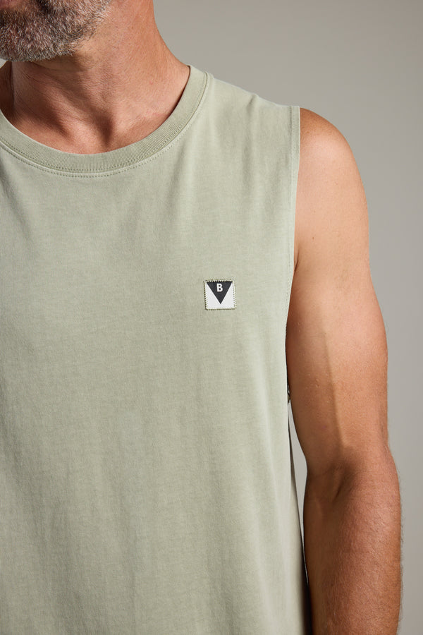 A man wears the Barkers Newton Washed Singlet, a sleeveless light green organic cotton top with a small black and white triangular chest logo. The photo features his upper torso and bearded face.
