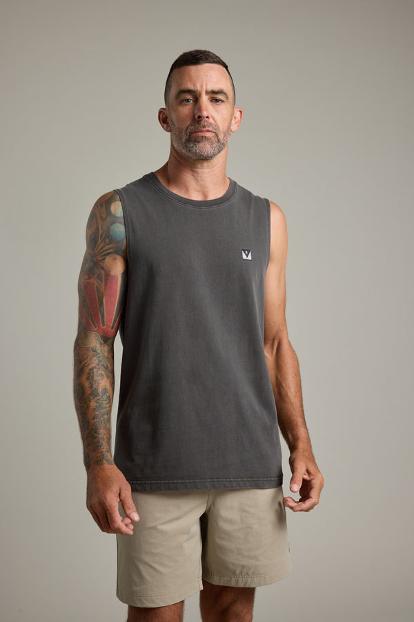 A man with short hair and a beard stands facing forward, wearing the Barkers Newton Washed Singlet in dark gray and light khaki shorts. His left arm's colorful tattoos stand out against the plain light gray background.