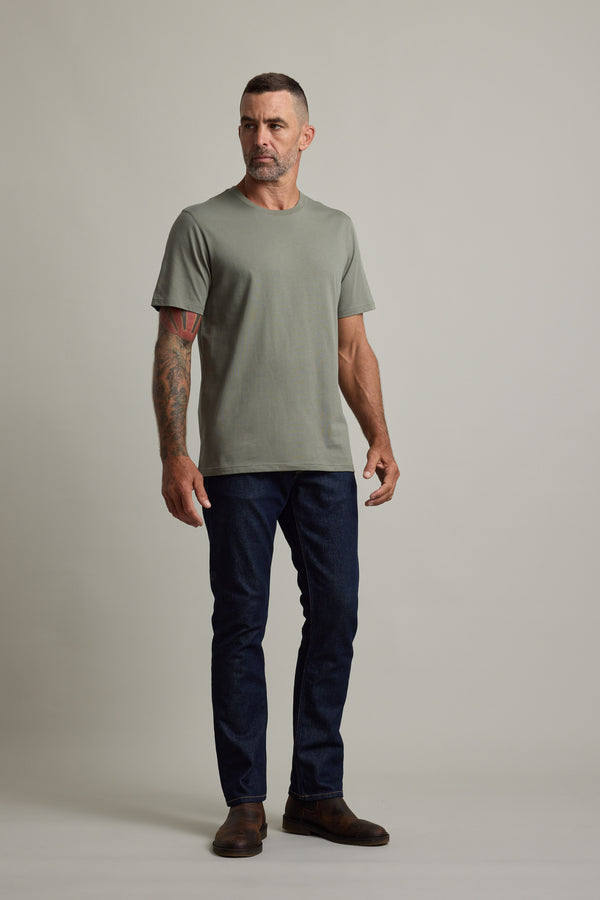 Tattooed man wears Barkers Good Earth Cotton Essential Tee, dark jeans, and brown boots.