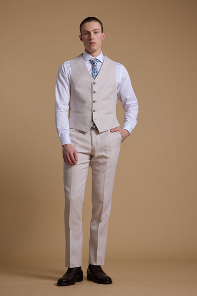 A man stands against a beige background wearing the Barkers Somerton Texture Waistcoat and matching pants, styled with a white shirt, blue patterned tie, and dark brown shoes—an ideal summer tailoring look. One hand is in his pocket as he looks at the camera.