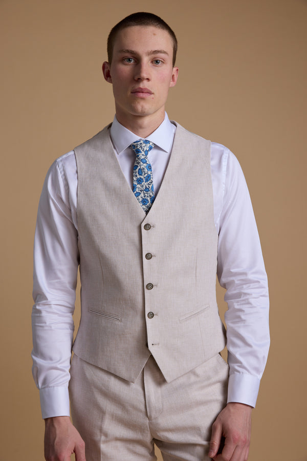 A man wears the Barkers Somerton Texture Waistcoat with matching pants and a white shirt paired with a blue floral tie, standing against a tan backdrop—an ideal look for summer tailoring.