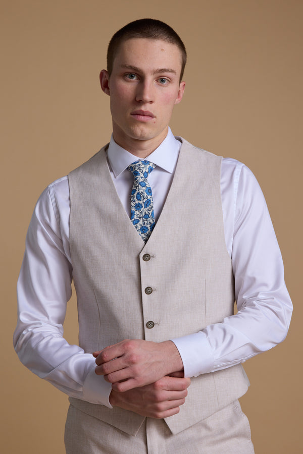 A young man with short hair models the Barkers Somerton Texture Waistcoat over a white dress shirt and blue floral tie, standing against a tan background with hands loosely clasped—a refined approach to summer tailoring.