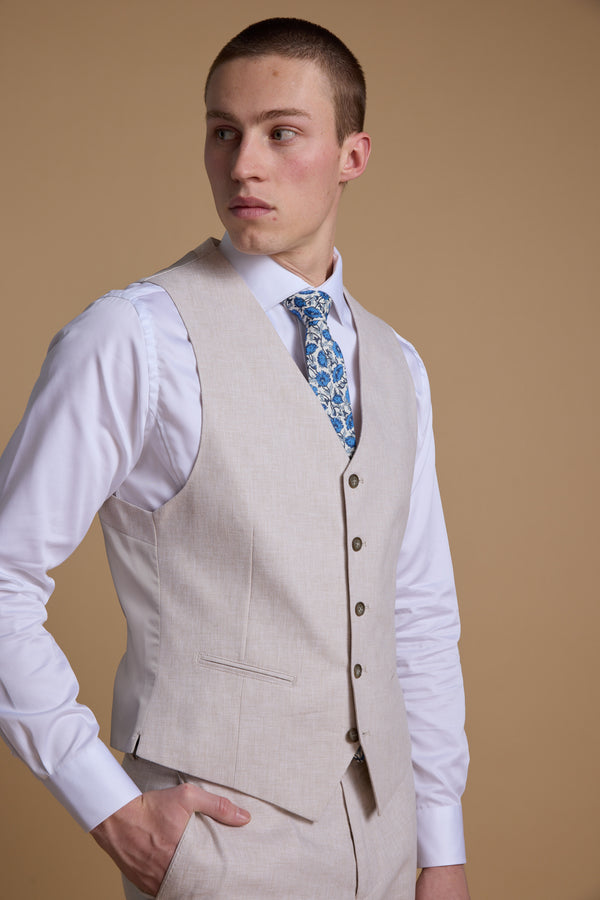 A young man models the Barkers Somerton Texture Waistcoat with matching trousers, a white shirt, and blue floral tie—effortless summer style against a tan backdrop.