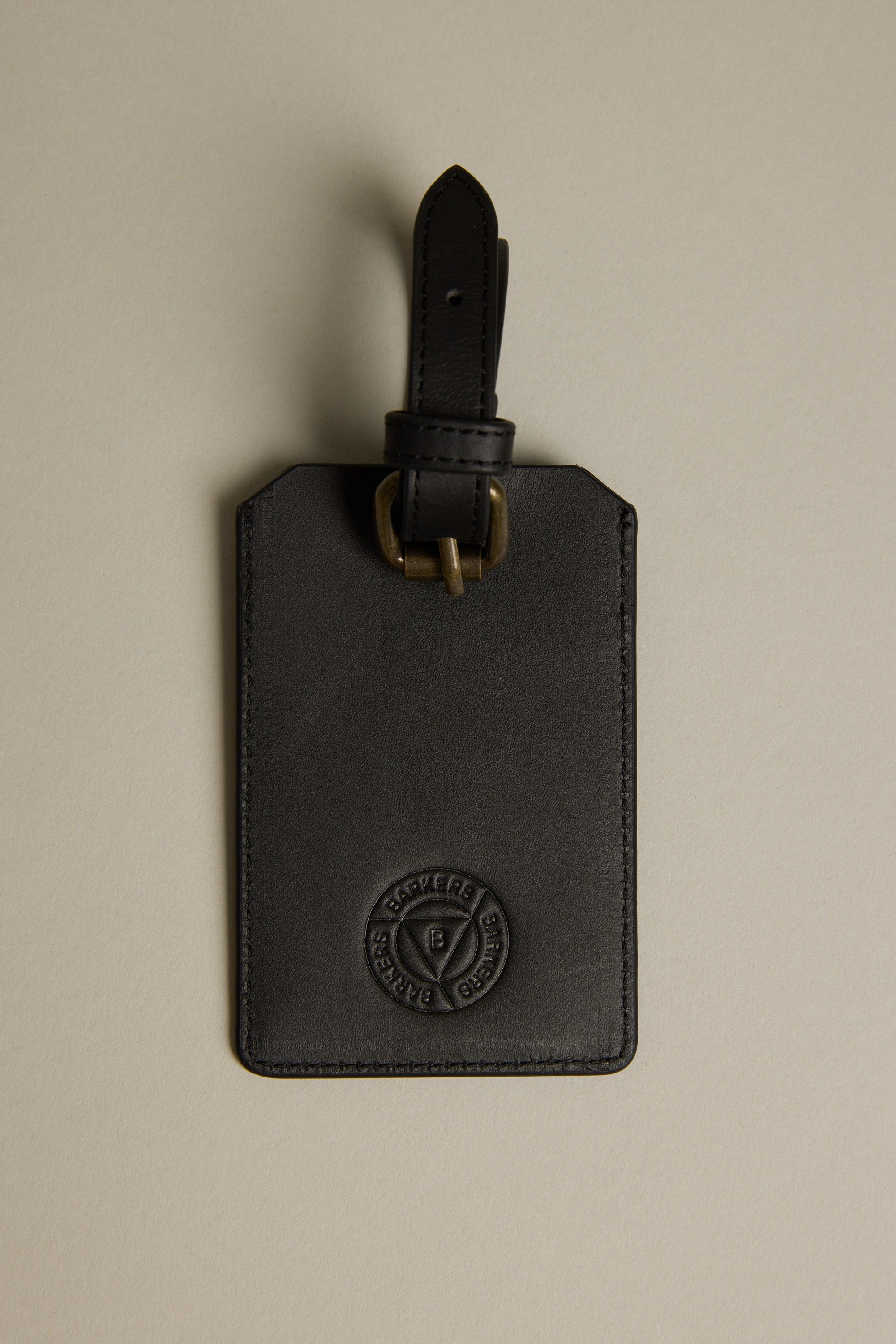The Barkers Voyage Leather Luggage Tag in black features a brass buckle strap and embossed circular logo, making it a stylish travel accessory or perfect corporate gift, shown here on a plain beige background.