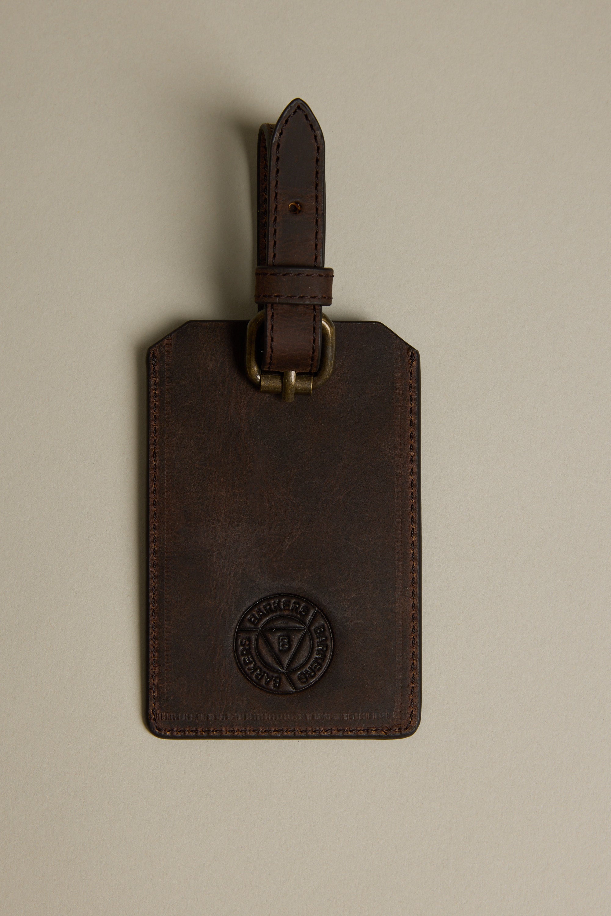 The Barkers Voyage Leather Luggage Tag, in brown with a brass buckle and embossed logo, is shown on a beige background. Visible stitching adds style—an ideal corporate gift for travelers.