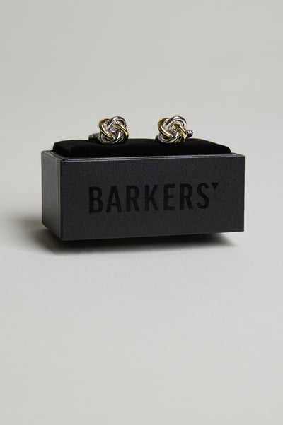 A pair of Barkers Knot Cufflink in silver and gold, elegantly displayed on a black velvet stand inside a sleek box with BARKERS branding. The perfect formal accessory for any special occasion.