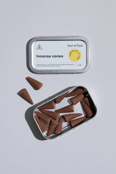 An open Earl of East EoE Jardin de La Lune Incense Cones tin sits on a light gray surface, showing several hand dipped cones inside and some tuberose-scented cones scattered nearby.