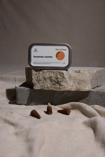 An Earl of East EoE Strand Incense Cones tin rests on stacked stone blocks, while five brown cones are arranged on textured beige fabric in the foreground.