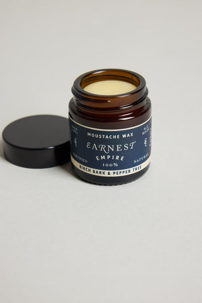 An open jar of Other Earnest Empire Moustache Wax with a dark lid beside it displays "Birch Bark & Pepper Tree" on the label. The light yellow wax inside is 100% natural and provides medium-to-firm hold for your style.