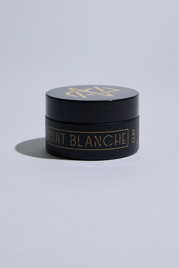 A black round container labeled "Nuit Blanche Clay" by Other in gold lettering sits on a light gray surface, casting a soft shadow. This matte finish hair clay is made in New Zealand and ideal for effortless styling.