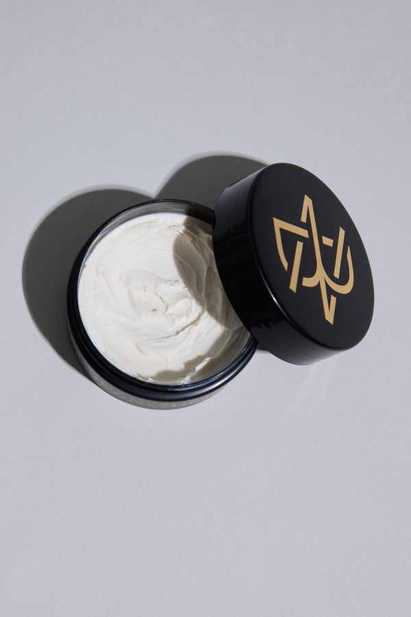 A black container of Other's Nuit Blanche Paste, featuring a gold geometric lid design and partially open to reveal smooth white styling paste inside, is displayed on a light gray surface.
