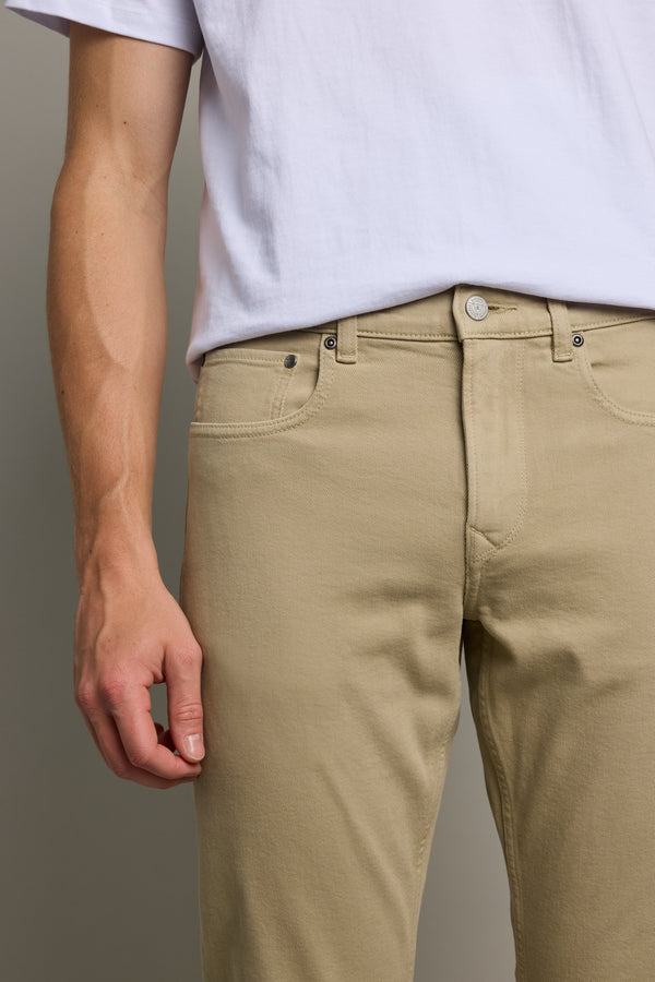 A person wearing a white t-shirt and Barkers Rambler Slim Coloured Jean in beige, shown from shoulders to thighs with only the left arm and hand visible, stands against a plain background.