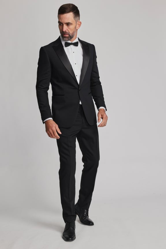 A man with short dark hair and a beard stands against a plain background, wearing a Barkers Black Bow Tie, white dress shirt, formal black tuxedo, and black dress shoes. He looks to the side with one hand by his waist.
