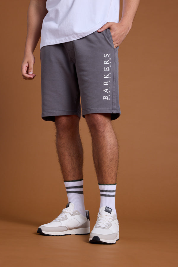 A person stands against a plain brown background wearing a white T-shirt, Barkers Organic Classic Print Track Short in grey with the heritage logo, white socks with black stripes, and white sneakers. The upper face and head are not visible.