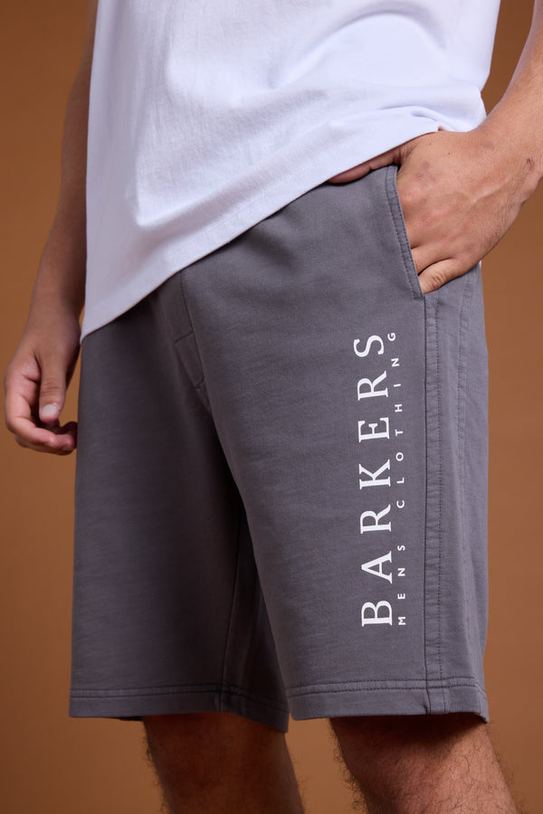 A person wears a white T-shirt and gray Barkers Organic Classic Print Track Short with one hand in a pocket. "BARKERS MENS CLOTHING" is printed vertically on the relaxed fit shorts. The background is brown.