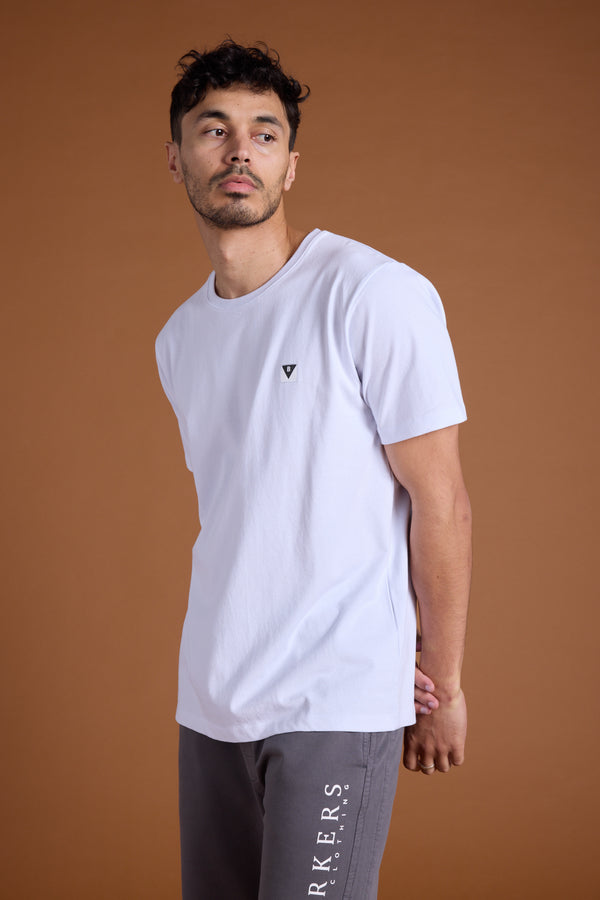 A man with short dark hair and facial hair wears the Barkers Newton Washed Tee, an oversized box fit organic cotton t-shirt, and gray sweatpants, standing against a solid brown background and looking slightly to his left.