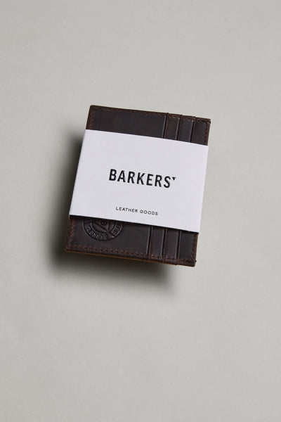 The Barkers Taylor Leather Card Wallet, a dark brown Leather Working Group certified slim card holder, is partially wrapped in a white paper band labeled BARKERS and displayed on a light grey surface.