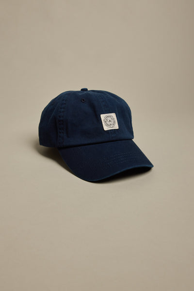 The Barkers Washed Cotton Cap by Barkers is a navy blue vintage wash hat with a curved brim and a small white rectangular patch featuring a circular logo on the front, shown against a plain beige background.